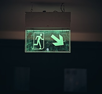A glowing green emergency exit sign features a person walking through a door with an arrow pointing downwards and to the right. The sign is enclosed in a rectangular box suspended from the ceiling by two wires against a dark background.
