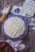 A homemade cake with textured frosting sits on a blue patterned plate. Surrounding items include scattered flour, a rolling pin, eggs, a handwritten recipe in a notebook, photographs, and a bowl of apples. The setting is rustic, with a wooden table surface.