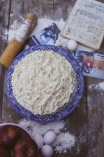 A homemade cake with textured frosting sits on a blue patterned plate. Surrounding items include scattered flour, a rolling pin, eggs, a handwritten recipe in a notebook, photographs, and a bowl of apples. The setting is rustic, with a wooden table surface.