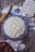 A homemade cake with textured frosting sits on a blue patterned plate. Surrounding items include scattered flour, a rolling pin, eggs, a handwritten recipe in a notebook, photographs, and a bowl of apples. The setting is rustic, with a wooden table surface.