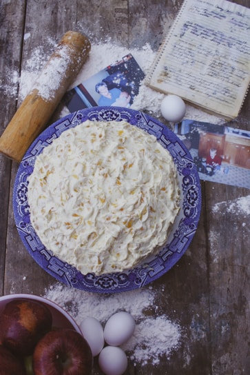 A homemade cake with textured frosting sits on a blue patterned plate. Surrounding items include scattered flour, a rolling pin, eggs, a handwritten recipe in a notebook, photographs, and a bowl of apples. The setting is rustic, with a wooden table surface.