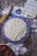 A homemade cake with textured frosting sits on a blue patterned plate. Surrounding items include scattered flour, a rolling pin, eggs, a handwritten recipe in a notebook, photographs, and a bowl of apples. The setting is rustic, with a wooden table surface.