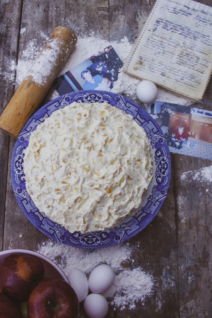 A homemade cake with textured frosting sits on a blue patterned plate. Surrounding items include scattered flour, a rolling pin, eggs, a handwritten recipe in a notebook, photographs, and a bowl of apples. The setting is rustic, with a wooden table surface.