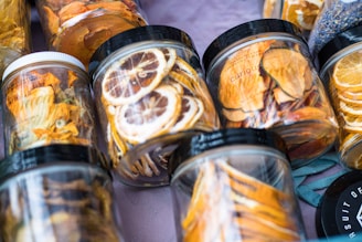 A variety of jars filled with dried fruits arranged on a surface. The jars contain slices of fruits such as lemons and oranges, with visible vibrant colors. Each jar has a black or white lid, and the setup gives a sense of freshness and a homemade touch.