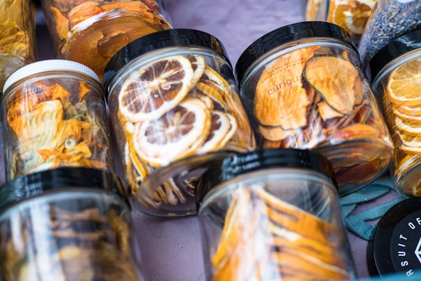 A variety of jars filled with dried fruits arranged on a surface. The jars contain slices of fruits such as lemons and oranges, with visible vibrant colors. Each jar has a black or white lid, and the setup gives a sense of freshness and a homemade touch.