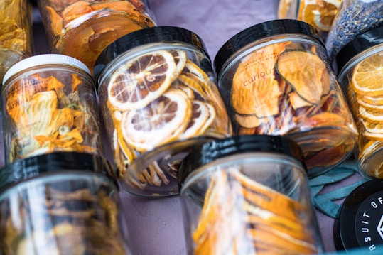 A variety of jars filled with dried fruits arranged on a surface. The jars contain slices of fruits such as lemons and oranges, with visible vibrant colors. Each jar has a black or white lid, and the setup gives a sense of freshness and a homemade touch.