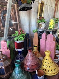 A display of decorative items including colorful ceramic tajines with intricate designs, various painted model structures resembling minarets or towers, ornate lanterns, and vibrant potted artificial plants with red berries and yellow flowers. The arrangement is set against a backdrop featuring mirrors and framed artworks.