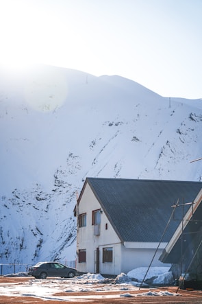 A snowy mountain landscape with a two-story building featuring a steep roof in the foreground. The sunlight is shining over the mountain, casting a bright glow, while a small car is parked next to the building on a snow-covered ground.