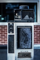 An old, industrial-looking communication device with the label 'Hamilton Tell-R-TV Integrated Audiovisual Communication System' is positioned in front of a brick wall. The device features buttons and a speaker, with visible wear and grime on its surface.