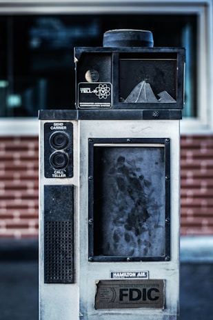 An old, industrial-looking communication device with the label 'Hamilton Tell-R-TV Integrated Audiovisual Communication System' is positioned in front of a brick wall. The device features buttons and a speaker, with visible wear and grime on its surface.