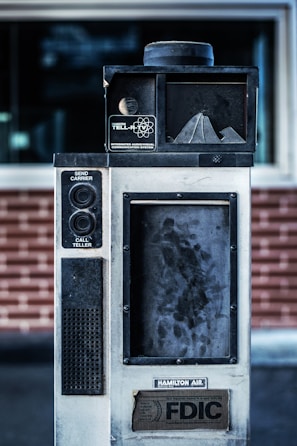 An old, industrial-looking communication device with the label 'Hamilton Tell-R-TV Integrated Audiovisual Communication System' is positioned in front of a brick wall. The device features buttons and a speaker, with visible wear and grime on its surface.