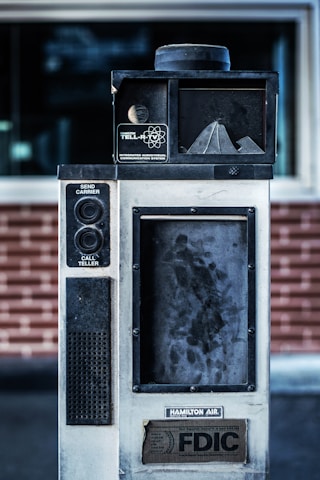 An old, industrial-looking communication device with the label 'Hamilton Tell-R-TV Integrated Audiovisual Communication System' is positioned in front of a brick wall. The device features buttons and a speaker, with visible wear and grime on its surface.