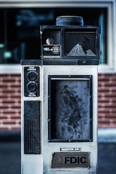 An old, industrial-looking communication device with the label 'Hamilton Tell-R-TV Integrated Audiovisual Communication System' is positioned in front of a brick wall. The device features buttons and a speaker, with visible wear and grime on its surface.
