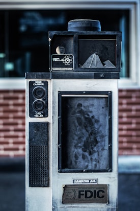 An old, industrial-looking communication device with the label 'Hamilton Tell-R-TV Integrated Audiovisual Communication System' is positioned in front of a brick wall. The device features buttons and a speaker, with visible wear and grime on its surface.