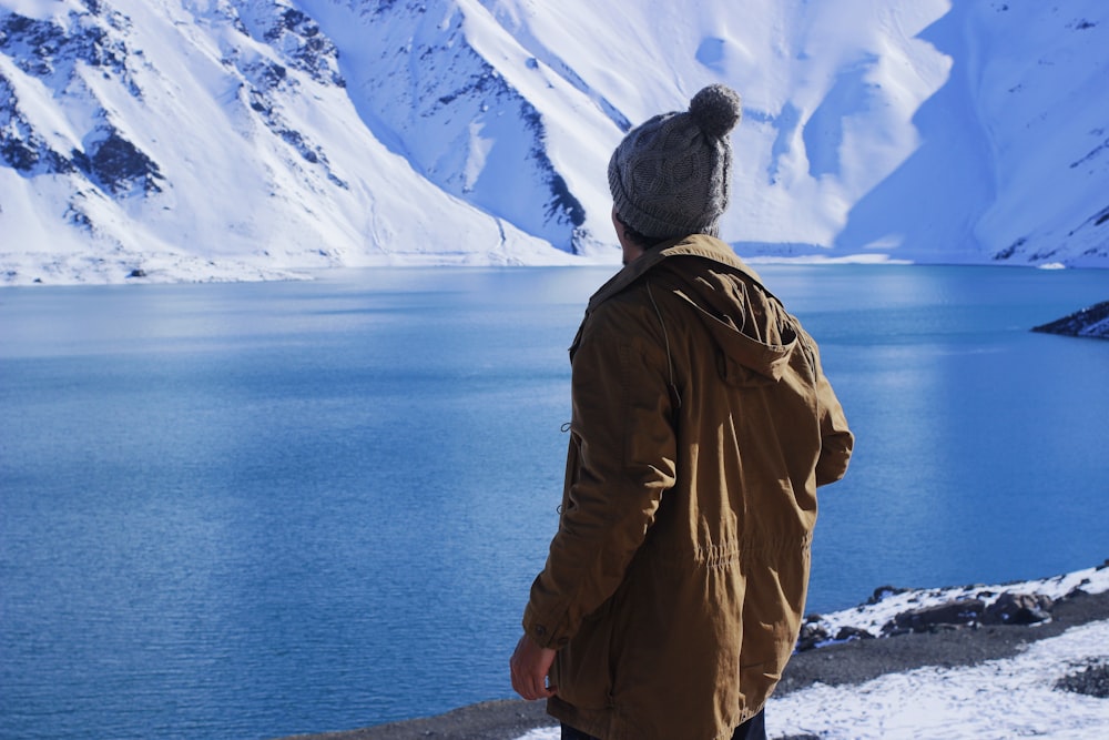 man in brown jacket standing on snow covered ground looking at blue sea during daytime