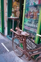 A creatively designed bicycle with an unusual metallic and mechanical structure is parked outside a shop. A skeleton is posed at a table next to the shop's entrance, which is highlighted with green paint. The window displays various bottles of absinthe and advertises the store as an absinthe bar.