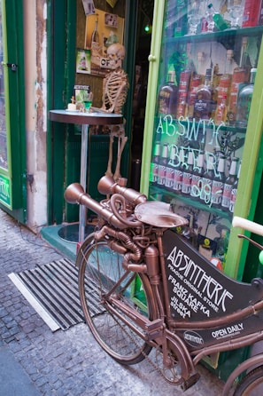A creatively designed bicycle with an unusual metallic and mechanical structure is parked outside a shop. A skeleton is posed at a table next to the shop's entrance, which is highlighted with green paint. The window displays various bottles of absinthe and advertises the store as an absinthe bar.