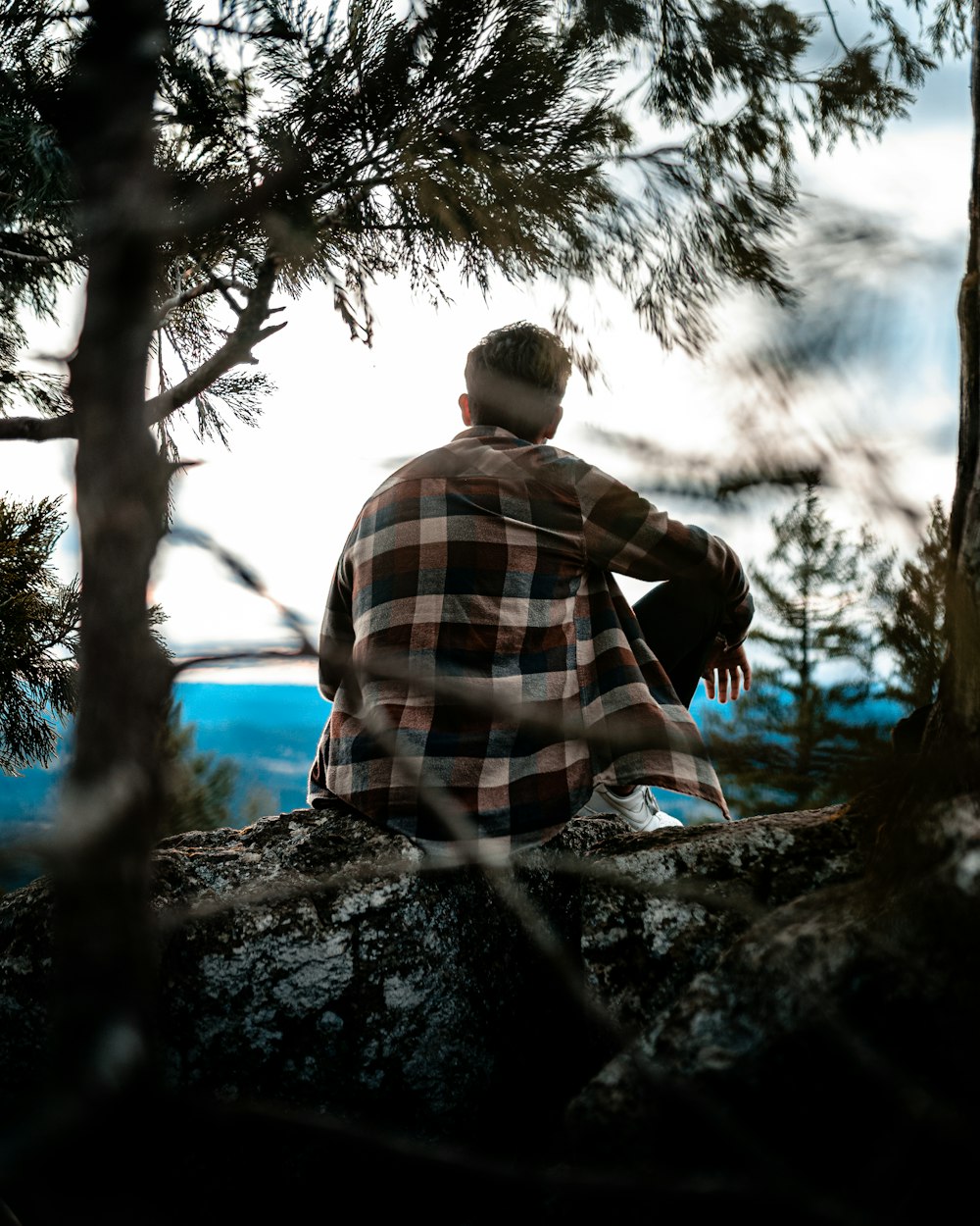 man in brown and blue plaid dress shirt sitting on rock near tree during daytime