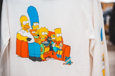 A printed image on a white shirt depicts a family of cartoon characters sitting on an orange couch, each character distinct in appearance and holding various items like a remote control, popcorn, and a drink. Surrounding them are several small objects on the floor, including a TV remote and a popcorn container.