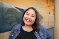 A smiling woman with shoulder-length dark hair and a black top is wearing a denim jacket. She is standing in front of a painted mural that seems to depict natural elements, primarily in shades of blue and yellow.
