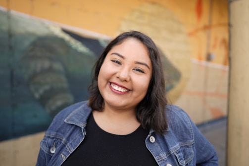 A smiling woman with shoulder-length dark hair and a black top is wearing a denim jacket. She is standing in front of a painted mural that seems to depict natural elements, primarily in shades of blue and yellow.