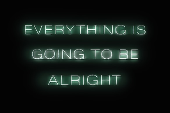 Neon green text on a black background reads 'EVERYTHING IS GOING TO BE ALRIGHT'. The letters are bright and evenly spaced, creating a calming and reassuring message.
