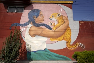 A mural depicts a woman with braided blue hair holding a baby jaguar. The woman is wearing a white top and blue-green skirt, and her arm features a tattoo. The background is a large circular design with a reddish-brown wall and some plants growing nearby.