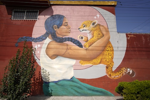 A mural depicts a woman with braided blue hair holding a baby jaguar. The woman is wearing a white top and blue-green skirt, and her arm features a tattoo. The background is a large circular design with a reddish-brown wall and some plants growing nearby.