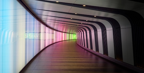 A modern, curving hallway with a futuristic design features illuminated walls displaying a gradient of colors ranging from blue to pink to green. The space has a sleek and minimalistic appearance with dark accents and a wooden floor.