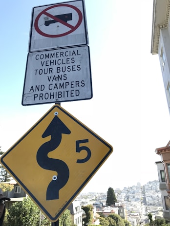 Two road signs are visible: a white rectangular sign with a symbol of a truck crossed out, indicating that commercial vehicles, tour buses, vans, and campers are prohibited, and a yellow diamond-shaped sign with a winding road symbol and the number 5, suggesting a sharp curve or winding road ahead.