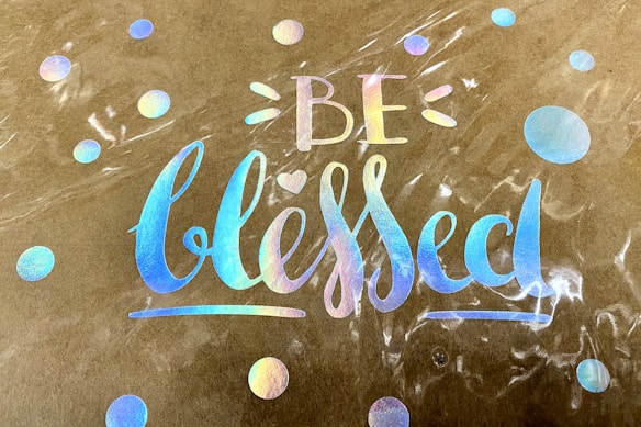 A decorative design features the words 'Be blessed' in iridescent rainbow lettering on a brown paper background. Surrounding the text are several circular dots also in a reflective rainbow pattern, giving a colorful and uplifting visual effect.