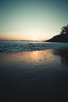 A tranquil beach scene at sunset with gentle waves lapping against the shore. The horizon is softly illuminated by the setting sun, casting a warm glow over the water. Silhouettes of cliffs and trees add contrast to the serene landscape.