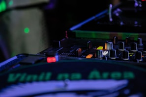 A DJ mixing console with various knobs, buttons, and sliders is illuminated under ambient lighting. A turntable with a vinyl showing the text 'Vinil na Área' is prominently visible. The scene suggests a live music performance setup.