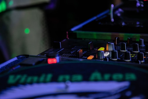 A DJ mixing console with various knobs, buttons, and sliders is illuminated under ambient lighting. A turntable with a vinyl showing the text 'Vinil na Área' is prominently visible. The scene suggests a live music performance setup.