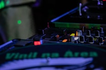 A DJ mixing console with various knobs, buttons, and sliders is illuminated under ambient lighting. A turntable with a vinyl showing the text 'Vinil na &Aacute;rea' is prominently visible. The scene suggests a live music performance setup.