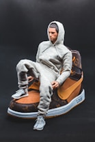A small figurine of a person wearing a gray tracksuit is sitting on a large sneaker. The sneaker has a luxurious design with brown and black tones, featuring a swoosh and patterns resembling those of a high-end brand. The background is a plain dark color, highlighting the subjects in the foreground.