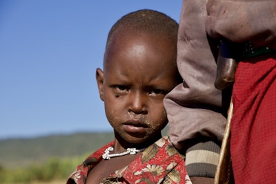 A young child wearing a patterned red shirt and a beaded necklace stands closely beside an adult. The child has a serious expression, and there is a natural landscape with a clear blue sky in the background.