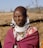 An individual wearing traditional attire, including a patterned shawl and an array of beaded jewelry, stands outside in a sunny setting. The background consists of natural elements such as dried grass and dirt, hinting at a rural environment.