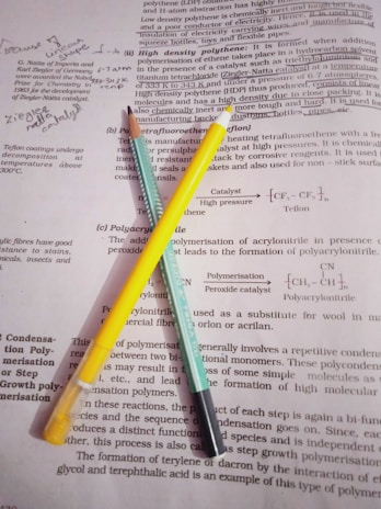 Two pencils, one yellow and one turquoise, are placed on top of pages containing printed text and handwritten notes. The text appears to be from a chemistry book or study material, discussing topics like polymerisation and catalysts. Handwritten annotations and diagrams are visible among the printed words.