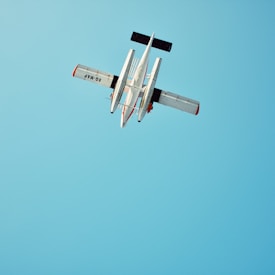 A small aircraft is captured mid-flight against a clear blue sky. The plane is viewed from below, showcasing its outstretched wings and tail with distinct red and white markings.