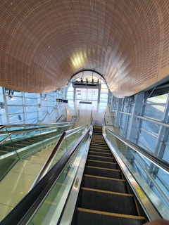 A modern, well-lit interior space featuring an escalator and a glass elevator. The architecture includes a curved wooden ceiling, extensive use of glass, and metal accents. The escalator descends towards sliding glass doors at the far end of the room, and there are glass walls on either side with views of the outside.