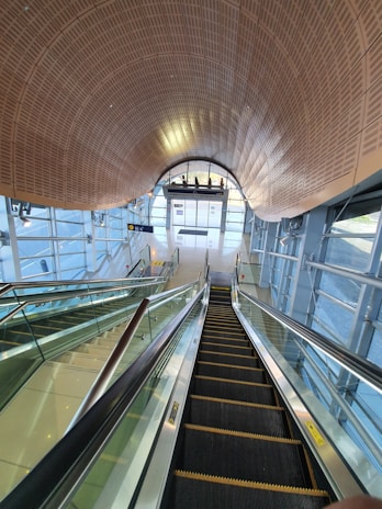 A modern, well-lit interior space featuring an escalator and a glass elevator. The architecture includes a curved wooden ceiling, extensive use of glass, and metal accents. The escalator descends towards sliding glass doors at the far end of the room, and there are glass walls on either side with views of the outside.