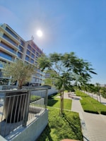 A modern multi-story apartment building stands against a clear blue sky with the sun shining brightly. In the foreground, landscaped greenery includes neatly trimmed hedges and several trees along a paved pathway. The building features balconies with glass railings, and there is a metal railing near a small tree planted in a raised bed.