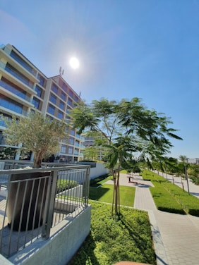 A modern multi-story apartment building stands against a clear blue sky with the sun shining brightly. In the foreground, landscaped greenery includes neatly trimmed hedges and several trees along a paved pathway. The building features balconies with glass railings, and there is a metal railing near a small tree planted in a raised bed.