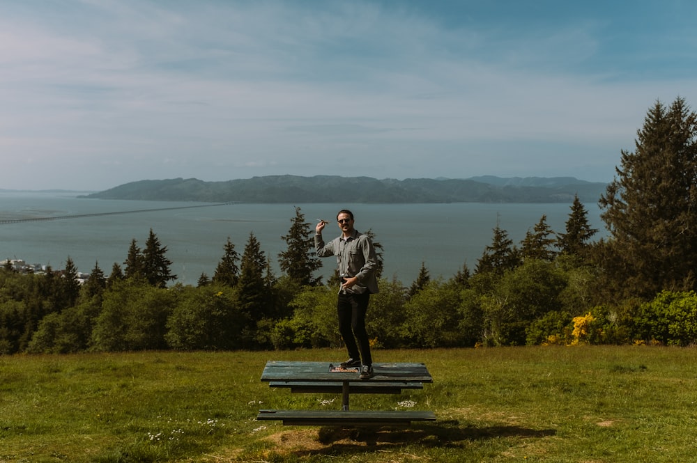 man in black jacket and blue denim jeans standing on blue wooden bench near green trees
