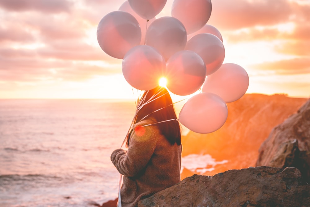 Mujer en chaqueta marrón sosteniendo globos en la playa durante la puesta del sol