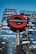 A prominent London Underground sign stands in the foreground against the backdrop of modern skyscrapers, with illuminated windows reflecting the evening sky.