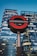 A prominent London Underground sign stands in the foreground against the backdrop of modern skyscrapers, with illuminated windows reflecting the evening sky.