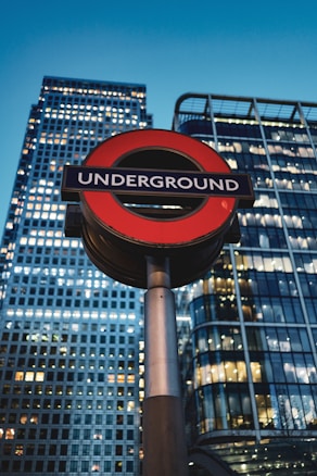 A prominent London Underground sign stands in the foreground against the backdrop of modern skyscrapers, with illuminated windows reflecting the evening sky.
