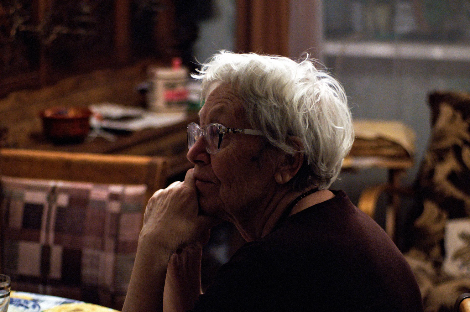 An older woman in fine rectangular reading glasses turning towards a window in a warm-walled room